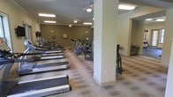 This fitness center offers a comprehensive workout environment designed to accommodate a range of exercise needs. The main workout area is generously sized, featuring a row of treadmills from brands including TRUE and Nautilus, alongside elliptical machines and stationary bikes arranged to maximize the use of space. Structural columns punctuate the floor plan, adding architectural character while delineating the workout zones. The flooring consists of a patterned carpet tile in neutral tones, providing both comfort and durability underfoot. Overhead, multiple ceiling fans with integrated lighting ensure consistent air circulation throughout the space, complementing the flush-mounted fluorescent lighting that keeps the area well-lit. The walls are finished in a warm olive-grey tone, contributing to a focused and motivating atmosphere. Crown molding along the ceiling adds a refined touch to the otherwise utilitarian setting. A wall clock is mounted prominently, allowing users to keep track of their workout time. An adjacent area visible through an open archway suggests additional amenities nearby, extending the overall utility of this single-floor facility.