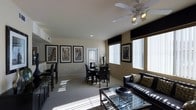 This single-floor space features an open-plan layout that seamlessly combines living and dining areas, creating a cohesive and functional environment. The interior is finished with neutral beige walls and light-toned carpet, providing a clean, versatile backdrop that complements the contemporary dark-toned furnishings throughout.

The living area is anchored by a sleek black sofa adorned with patterned accent pillows, positioned alongside a glass-top coffee table that adds a reflective, modern touch. A ceiling fan with integrated lighting is centrally mounted overhead, contributing both practical airflow and ambient illumination.

The dining area features a round dark-wood table surrounded by matching chairs, accented by a decorative vase arrangement at its center. Large windows fitted with white vertical blinds and dark valances allow generous natural light to filter into the space, casting bright, patterned sunlight across the floor.

A collection of framed architectural photography lines the walls, lending an urban, sophisticated character to the space. A partial view of a kitchen counter with bar seating is visible to one side, suggesting additional functionality. A door at the far end of the dining area provides access to an exterior or adjacent area.