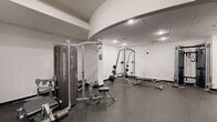Located at 500 Merrimac Trail in Williamsburg, Virginia, this two-floor fitness facility offers a well-appointed exercise environment spread across two distinct levels. The upper floor serves as the primary cardio and functional training area, featuring rubber flooring throughout, bright white walls, and a distinctive tray ceiling with blue accents and recessed lighting. Large windows along one wall allow natural light to fill the space, creating an open and energizing atmosphere. This level accommodates a row of elliptical trainers and stationary bikes, a dumbbell rack with an adjustable bench, and a dedicated functional training room equipped with stability balls, medicine balls, and a wall-mounted mat — ideal for stretching and bodyweight exercises. A staircase with dark wood railings and a vibrant yellow-green accent wall connects the two floors, adding a pop of color to the transition space. The lower floor features a curved architectural ceiling detail that lends visual interest to the strength training area. This level houses a multi-station cable machine, a power rack with bench and free weights, and additional strength equipment, all set on durable rubber flooring with recessed and drop-ceiling lighting. Together, the two floors provide a comprehensive and well-equipped fitness facility suited for a full range of workout routines.