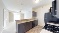 This single-floor residence offers a thoughtfully designed open-concept layout that seamlessly connects the kitchen and living areas. The kitchen is equipped with rich, dark espresso-toned cabinetry that provides ample storage, complemented by granite-style countertops that add a touch of elegance. A stainless steel sink is centrally positioned along the counter, flanked by a black dishwasher and refrigerator, giving the kitchen a sleek, cohesive look. Stylish pendant lights hang above the kitchen counter, providing focused task lighting while enhancing the overall aesthetic. The open pass-through design allows for easy interaction between the kitchen and the adjoining living space, which features a ceiling fan and large window dressed with vertical blinds, allowing natural light to filter in and brighten the interior. The flooring transitions from tile in the kitchen to carpet in the living area, clearly defining each functional zone. The textured ceiling adds subtle visual interest throughout. Overall, the space presents a clean, functional, and modern ambiance well-suited for comfortable everyday living.