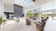 Located at 301 Commons Park South in Stamford, Connecticut, this generously sized amenity lounge is a standout communal space defined by its dramatic architectural design and upscale finishes. The single-floor space is anchored by soaring double-height ceilings and a large skylight that floods the interior with abundant natural light throughout the day. Floor-to-ceiling windows and glass doors line the perimeter, offering sweeping views of the surrounding greenery and providing seamless visual connectivity to the outdoors.

The open layout is thoughtfully organized into distinct functional zones. A comfortable lounge area features plush seating arrangements with neutral-toned armchairs and sofas centered around a sleek linear fireplace set against a bold charcoal accent wall. Adjacent to this is a game area complete with a billiards table, as well as a dining zone with a solid wood table and upholstered seating. A vibrant nook with a tufted gold banquette adds a pop of color near the entryway.

The fully equipped kitchen showcases white cabinetry, granite countertops, stainless steel appliances, and a curved island with bar seating, complemented by sculptural pendant lighting. A grab-and-go retail display adds convenience. Light herringbone-patterned flooring unifies the entire space, contributing to its clean, contemporary aesthetic.