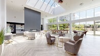 Located at 301 Commons Park South in Stamford, Connecticut, this generously sized amenity lounge is a standout communal space defined by its dramatic architectural design and upscale finishes. The single-floor space is anchored by soaring double-height ceilings and a large skylight that floods the interior with abundant natural light throughout the day. Floor-to-ceiling windows and glass doors line the perimeter, offering sweeping views of the surrounding greenery and providing seamless visual connectivity to the outdoors.

The open layout is thoughtfully organized into distinct functional zones. A comfortable lounge area features plush seating arrangements with neutral-toned armchairs and sofas centered around a sleek linear fireplace set against a bold charcoal accent wall. Adjacent to this is a game area complete with a billiards table, as well as a dining zone with a solid wood table and upholstered seating. A vibrant nook with a tufted gold banquette adds a pop of color near the entryway.

The fully equipped kitchen showcases white cabinetry, granite countertops, stainless steel appliances, and a curved island with bar seating, complemented by sculptural pendant lighting. A grab-and-go retail display adds convenience. Light herringbone-patterned flooring unifies the entire space, contributing to its clean, contemporary aesthetic.