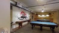 This single-floor recreation space is purpose-built for social entertainment, offering a well-appointed game room atmosphere. The centerpiece of the room includes multiple full-sized pool tables with rich dark wood frames and vibrant blue felt surfaces, each illuminated by elegant pendant light fixtures that cast focused, warm light over the playing areas. A full-length shuffleboard table lines one wall, providing an additional classic game option for guests. The space features an exposed industrial ceiling with visible ductwork and open structural elements, lending an urban, loft-like character to the room. A bold, eye-catching mural on the wall depicts a stylized city skyline accented with dramatic red and grey graphic elements, serving as a strong visual focal point. The flooring is covered in a neutral carpet, contributing to the room's relaxed and comfortable ambiance. Cabinetry along the back wall suggests additional storage or service functionality. The overall design blends industrial aesthetics with recreational comfort, making it a versatile and inviting gathering space suited for casual social interaction and organized game play alike.