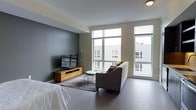 This single-floor space showcases a clean, modern design that seamlessly integrates the living and sleeping areas into one cohesive open layout. The interior is defined by a neutral color palette, with warm beige walls contrasted by a bold slate-gray accent wall that anchors the living zone and adds visual depth to the space.

Large floor-to-ceiling windows dominate one wall, flooding the interior with abundant natural light and providing open views of the surrounding urban streetscape. The windows are equipped with roller shades for adjustable privacy and light control.

Dark-toned hardwood flooring runs throughout, lending warmth and continuity across the entire space. A charcoal-upholstered sofa is positioned to face a wall-mounted flat-screen television, which sits atop a natural wood media console — a detail that introduces organic warmth against the otherwise contemporary backdrop. A minimalist coffee table and a textured area rug in dark tones define the seating arrangement.

The sleeping area flows naturally from the living space, maintaining the open-plan character of the unit. Flush-mount ceiling lighting provides practical, unobtrusive illumination throughout, completing the streamlined, functional aesthetic of this well-appointed space.