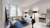 This thoughtfully designed single-floor space exudes understated elegance through its carefully curated interiors and classic architectural elements. The living area serves as the heart of the space, highlighted by tall, multi-pane sash windows that flood the room with abundant natural light and offer a glimpse of lush greenery beyond. Crisp white crown molding frames the ceiling, adding a refined architectural touch that complements the warm, neutral wall tones.

The living room is furnished with a clean-lined white sofa accented by deep burgundy cushions, paired with contemporary tufted armchairs and a rustic wood coffee table atop a rich navy blue area rug — a combination that balances classic and modern sensibilities. Flanking the seating area, tall black floor lamps with structured shades provide warm ambient lighting, while a distinctive circular metal wall art piece serves as a bold focal point. A partially visible dining area with a round white table and an oval-backed chair hints at seamless transitions between living and dining spaces.

The light wood flooring throughout enhances the sense of openness and warmth. Overall, the space presents a cohesive, sophisticated atmosphere ideal for both relaxed living and refined entertaining.