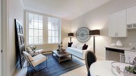 This thoughtfully designed single-floor space exudes understated elegance through its carefully curated interiors and classic architectural elements. The living area serves as the heart of the space, highlighted by tall, multi-pane sash windows that flood the room with abundant natural light and offer a glimpse of lush greenery beyond. Crisp white crown molding frames the ceiling, adding a refined architectural touch that complements the warm, neutral wall tones.

The living room is furnished with a clean-lined white sofa accented by deep burgundy cushions, paired with contemporary tufted armchairs and a rustic wood coffee table atop a rich navy blue area rug — a combination that balances classic and modern sensibilities. Flanking the seating area, tall black floor lamps with structured shades provide warm ambient lighting, while a distinctive circular metal wall art piece serves as a bold focal point. A partially visible dining area with a round white table and an oval-backed chair hints at seamless transitions between living and dining spaces.

The light wood flooring throughout enhances the sense of openness and warmth. Overall, the space presents a cohesive, sophisticated atmosphere ideal for both relaxed living and refined entertaining.