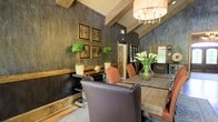 This single-floor space showcases a striking rustic-modern aesthetic that masterfully blends natural materials with refined design elements. The interior is defined by dramatic dark, textured walls that provide a rich backdrop throughout, complemented by exposed wooden ceiling beams that add architectural character and a sense of warmth. Natural wood tones are carried throughout the space, from the sturdy reclaimed wood dining table to the wainscoting detail along the lower walls, creating a cohesive and grounded feel. The dining area serves as a centerpiece, furnished with a substantial rustic wood table surrounded by a mix of tufted leather and upholstered chairs in warm terracotta and chocolate brown tones, evoking a sense of casual luxury. Overhead, an elegant drum chandelier with multiple candelabra bulbs provides ambient lighting that enhances the inviting atmosphere. Decorative elements are thoughtfully curated, including a gallery wall of ornate sunburst-style framed artwork, potted topiary plants, and a fresh floral arrangement that bring life and texture to the space. Natural light filters in through windows visible in the background, softening the deep tones of the interior. Overall, the space exudes a warm, upscale ambiance ideal for gathering and entertaining.