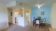 Chestnut Ridge Apartments presents a thoughtfully designed single-floor residence that blends functionality with a welcoming ambiance. The open-concept layout connects the kitchen and dining area seamlessly, creating an efficient and social living environment. The kitchen features light wood cabinetry, white appliances including a range and refrigerator, and tile flooring, providing ample storage and workspace for everyday meal preparation. A pass-through peninsula separates the kitchen from the adjacent spaces, adding a sense of openness while maintaining distinct zones. The dining area is accented by a bold blue feature wall that adds a pop of color and character to the space, complemented by a ceiling fan overhead and plush carpeted flooring underfoot. The neutral wall tones throughout balance the vibrant accent wall, creating a cohesive and inviting atmosphere. A textured ceiling adds a subtle design detail across the unit. The layout makes practical use of every square foot, offering a comfortable and functional home within the Chestnut Ridge Apartments community.