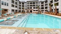 This residential community is centered around a striking courtyard designed for outdoor living and leisure. The focal point is a generously sized swimming pool with crystal-clear turquoise water, framed by a broad, light-toned tile and herringbone-patterned brick pool deck. The deck is well-appointed with numerous lounge chairs accented in teal, accompanied by matching market umbrellas, creating a cohesive and inviting aesthetic throughout the outdoor space.

The surrounding multi-story building features a contemporary architectural design, combining gray horizontal siding, white brick detailing, and dark-trimmed windows and balconies that overlook the courtyard. Tall palm trees are strategically planted throughout, lending a coastal, resort-like ambiance to the environment.

A covered pavilion with an outdoor seating area is situated at the far end of the pool, providing a shaded gathering space. The main building entrance is distinguished by a grand, covered entry portico supported by substantial columns, with wide glass doors framed in dark metal and flanked by large decorative planters. Herringbone brick paving continues from the pool deck to the building entrance, creating a seamless and polished hardscape design that ties the entire outdoor area together cohesively.