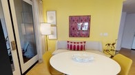 This single-floor space makes a striking first impression with its bold, sunny yellow walls that immediately set a warm and energetic tone. The dining area is a standout feature, centered around a sleek white round table paired with mustard yellow chairs that complement the room's vibrant color palette. A cozy upholstered bench with a bold patterned throw pillow in pink and cream adds both comfort and style to the seating arrangement. The walls are thoughtfully curated with eclectic artwork, including a eye-catching red and pink geometric patterned framed piece that incorporates a mirror element, alongside a pair of delicate botanical prints in neutral frames. Natural light filters in through sheer white curtains framed by light drapes, softening the brightness of the walls while maintaining an airy feel. A tall floor lamp with a classic white drum shade provides warm supplemental lighting. The hardwood flooring grounds the space with a natural warmth that balances the vivid color choices above. Overall, the space exudes a playful yet curated personality, blending retro-inspired design elements with modern furnishings for an atmosphere that is both lively and welcoming.