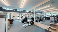 Located at 2260 Ringling Blvd in Sarasota, Florida, this contemporary fitness center is designed to deliver a premium workout experience in a thoughtfully planned single-floor space. The facility is characterized by soaring double-height ceilings accented with sleek linear LED lighting, creating a bright and energizing atmosphere throughout.

Large floor-to-ceiling windows line the perimeter, flooding the space with abundant natural light and offering views of the surrounding palm-lined landscape — a distinctly Floridian backdrop. The interior features a modern color palette of crisp white walls, deep teal accent panels, and cool blue-gray carpet flooring, lending the space a clean, professional aesthetic.

The cardio zone is generously equipped with multiple treadmills, elliptical trainers, stationary bikes, and rowing machines — many featuring integrated digital displays. Full-length mirrors are strategically positioned along the walls to enhance the sense of space and allow users to monitor their form. The strength and resistance training area offers a robust selection of Life Fitness cable machines, selectorized equipment, and a dedicated free weights section with a full dumbbell rack. A water station is conveniently accessible within the space. The overall layout promotes ease of movement, making this facility a well-rounded and inviting destination for fitness.