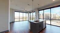 Located at 3233 Harmon Ave in Austin, TX, this single-floor residence showcases a sleek, contemporary design defined by dramatic floor-to-ceiling windows that flood the interior with natural light and frame sweeping views of the surrounding Austin skyline and lush treetops. Rich hardwood flooring flows throughout the open-concept main living area, creating a warm and cohesive aesthetic. The kitchen is a standout feature, boasting a large center island with quartz countertops, a deep single-basin sink with an industrial-style faucet, and stylish pendant lighting overhead. Cabinetry in a sophisticated gray tone pairs elegantly with stainless steel appliances, including a built-in microwave, range, and refrigerator, all set against a crisp white tile backsplash. The living area is generously proportioned, bathed in light from expansive windows, and finished with a ceiling fan for comfort. Bedrooms feature plush carpeting and their own floor-to-ceiling windows with ceiling fans, offering continued views of the cityscape. The bathrooms are finished with large-format tile, soaking tubs with tiled surrounds, and clean white vanities with marble-look countertops. A walk-in closet with built-in shelving and hanging rods adds practical storage. The overall ambiance is bright, modern, and urban.