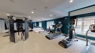 Located at 1177 Post Rd E in Westport, Connecticut, this professional fitness center is designed to support a comprehensive range of workout routines. The space is defined by bold teal accent walls contrasted against crisp white ceilings and neutral-toned flooring, creating an energizing yet polished atmosphere throughout.

The cardio zone lines the windowed perimeter, housing a row of treadmills, elliptical trainers, a stair climber, and a rowing machine — each equipped with individual digital displays. Natural light filters in through multiple windows, enhancing the open and motivating environment.

The strength training area is equally well-appointed, featuring a multi-functional RAX cable and functional training rig, a punching bag, medicine balls, battle ropes, and a full rack of dumbbells. A dedicated adjustable weight bench provides additional versatility for free weight exercises. A large wall mirror in the strength zone allows users to monitor form and visually expands the space.

A padded foam mat area offers space for stretching, floor work, and bodyweight training. The facility also includes a glass-partitioned room — suitable for private training or cool-down — along with built-in open shelving for storage, two private rooms accessed via white paneled doors, and recessed lighting throughout that ensures the space is consistently and evenly illuminated.
