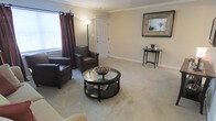 This two-story home offers a thoughtfully designed interior that balances comfort and classic style. The living room sets the tone for the space, showcasing neutral, warm-toned walls complemented by light beige carpeting that extends throughout. Crown molding adds a refined architectural detail, lending the room a polished, traditional character. Natural light filters in through a window dressed with rich, deep-burgundy curtains, creating a warm and inviting atmosphere. The room is well-proportioned, providing ample space for seating and relaxed gatherings. A circular glass-top coffee table serves as a central focal point, while a sleek floor lamp adds ambient lighting to the corner. A decorative framed mirror adorns one wall, enhancing the sense of depth and brightness in the room. A slim accent table nearby adds a subtle decorative touch. A clean white interior door completes the living area, maintaining the home's crisp, classic aesthetic. The overall ambiance throughout is cozy yet elegant, with a neutral palette that offers a versatile backdrop for a variety of décor styles. Spread across two floors, this home provides generous living space suited for comfortable everyday living.