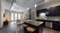 This single-floor residence showcases a contemporary urban design, blending industrial and modern elements throughout its open layout. The kitchen serves as a central focal point, featuring dark espresso cabinetry extending to the ceiling, a textured tile backsplash, stainless steel appliances, and a substantial granite-topped island with bar seating — ideal for both meal prep and casual dining. Track lighting and elegant pendant fixtures illuminate the space from above, while exposed ductwork along the ceiling adds an industrial character.

The open-concept design flows seamlessly into the living area, which is anchored by dark hardwood flooring and accented by soft, neutral-toned walls. A pair of French doors floods the room with natural light and provides outdoor access. The living room features elevated ceiling height, a ceiling fan, and comfortable furnishings that complement the overall aesthetic.

The bedroom is a calm retreat, with a ceiling fan, a large window dressed with blinds and curtains, dark hardwood flooring, and a warm, muted color palette. A gallery-style wall arrangement adds a curated touch to the space.

The bathroom continues the dark-cabinetry theme with a granite vanity countertop, an undermount sink, a large mirror, and tile flooring, offering a clean and functional finish throughout.