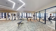Located at 5475 N University in Lauderhill, Florida, this expansive fitness center spans a generous single floor, offering an impressive and well-equipped workout facility. The space is defined by its striking architectural ceiling design, featuring geometric LED light fixtures that create a clean, modern ambiance while providing ample, evenly distributed illumination throughout.

The fitness center is divided into distinct functional zones. The cardio area is well-stocked with treadmills, ellipticals, a StairMaster, stationary bikes, and rowing machines, all positioned along large windows that allow natural light to filter in and provide views of the surrounding greenery. A vibrant motivational mural adds an energetic focal point to this section, complemented by a mounted flat-screen television.

The strength training area is equally comprehensive, housing a broad selection of cable machines, multi-station weight machines, free weight benches, and a full dumbbell rack. Floor-to-ceiling mirrors line the walls, visually expanding the space and allowing users to monitor their form. The patterned carpet tile flooring throughout provides both comfort underfoot and a polished aesthetic. Built-in shelving offers additional storage and organizational convenience. The overall layout is open, uncluttered, and thoughtfully designed to accommodate a wide range of fitness activities.