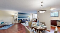 This single-floor, 2-bedroom, 1-bathroom unit is thoughtfully designed to maximize its intimate footprint. The open-plan living and dining area serves as the heart of the home, where rich hardwood-style flooring runs continuously underfoot. A bold blue-and-white horizontally striped accent wall adds a striking visual anchor to the living space, which benefits from a large window and sliding glass door that invite natural light and provide outdoor views of surrounding greenery. A pendant light fixture overhead defines the dining area, creating a natural separation within the open layout.

The kitchen features dark espresso cabinetry, white countertops, stainless steel appliances including a refrigerator and dishwasher, and a window that further enhances natural light. A hallway connects the main living area to the private quarters, lined with panel-style doors and warm flooring. The primary bedroom is a well-proportioned retreat with a closet, natural window views, and hardwood-style floors. The bathroom is finished with a dark vanity, white countertop, and a tub-shower combination. Storage is addressed throughout via dedicated closet space in the bedrooms.