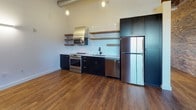 Located at 220 Merrimack in Lawrence, Massachusetts, this thoughtfully converted industrial unit masterfully blends historic character with modern finishes. The open-plan living area commands attention with soaring ceilings accented by original wood planking and exposed ductwork, while rich hardwood floors run throughout, grounding the space with warmth. Original exposed brick walls on multiple sides preserve the building's storied mill heritage and lend an authentic, textural depth to the interior.

The kitchen is a standout feature, outfitted with dark navy cabinetry, open wood-accented shelving, stainless steel appliances including a range with a professional-grade hood, dishwasher, and refrigerator, all set against a clean white tile backsplash. Pendant lighting adds an industrial-chic touch overhead.

Large arched windows allow natural light to pour into the living space, emphasizing the generous ceiling height and open feel. A convenient in-unit washer and dryer closet and additional storage are accessible through statement double doors set within the brick wall.

The bathroom is elegantly appointed with hexagonal patterned floor tile, subway-tiled walls, a wood-vanity with vessel sink and brass fixtures, and a separate shower — a refined contrast to the raw industrial aesthetic of the main living areas.