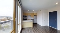 This single-floor, one-bedroom, one-bathroom unit offers a well-designed layout that maximizes its compact footprint. The open-concept living and kitchen area serves as the heart of the space, creating a seamless flow between cooking and lounging areas.

The kitchen is efficiently arranged with light maple cabinetry providing ample storage, a white countertop peninsula that doubles as a breakfast bar or prep space, and stainless steel appliances including a refrigerator, range, and microwave. Recessed lighting illuminates the kitchen area, while a single-basin sink sits neatly beneath upper cabinetry.

Hardwood-style flooring runs throughout the main living area, adding warmth and continuity to the space. Large floor-to-ceiling windows flood the interior with natural light, enhancing the sense of openness despite the unit's modest size. A sliding door element frames the window wall, providing a modern architectural detail.

White walls and clean lines maintain a neutral, contemporary aesthetic throughout. A separate door visible from the main area leads to the bedroom and bathroom, offering privacy and clear spatial delineation within the unit.