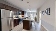 This single-floor residence showcases a thoughtfully designed open-concept layout with contemporary finishes and a cohesive neutral color palette. Light hardwood-style flooring flows throughout the main living areas, complementing the warm, modern aesthetic.

The kitchen serves as a central focal point, featuring rich dark cabinetry, granite countertops, and a full suite of stainless steel appliances. A kitchen island with pendant lighting overhead provides additional prep space and seating, seamlessly connecting the kitchen to the rest of the living area.

Adjacent to the kitchen, the dining area offers an intimate setting beneath a chandelier, with a round mirror accent and direct proximity to the entry door. The living room is bright and welcoming, with large floor-to-ceiling windows and a glass door leading to a private balcony, filling the space with natural light.

The bedroom continues the light, airy feel with oversized windows offering balcony views and linen drape curtains framing the natural light. Light wood flooring adds warmth to the room.

The bathroom features large-format tile flooring, a dark vanity with granite countertop, and direct access to a dedicated walk-in closet with built-in shelving and hanging space, providing ample storage throughout.