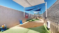 Located at 678 Bellflower Ave in Sunnyvale, California, this single-level outdoor play yard is thoughtfully designed to provide a safe and engaging environment for children. The space is enclosed by sturdy stucco perimeter walls and a natural wood picket fence, offering both security and a welcoming aesthetic.

Overhead, a series of large triangular shade sails — in both blue and white — are strategically positioned throughout the yard, providing generous coverage from the California sun while maintaining an open-air feel. String lights are strung across portions of the space, adding a warm, inviting ambiance.

The ground surface is a combination of artificial turf sections and packed natural terrain, creating distinct activity zones. One area features a colorful ball pit and a small wooden play structure with a striped canopy, designed for younger children. The perimeter wall is adorned with a charming white tree mural, adding a decorative and imaginative touch to the environment.

Additional amenities include a mounted interactive activity board in red on the wall, a water station, a wooden bench for supervision, and potted plants that introduce natural greenery. A small storage shed is also visible at one end of the yard.

Overall, the space is bright, well-organized, and purposefully arranged to support active, imaginative outdoor play in a secure and shaded setting.