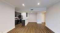 This single-floor residence offers a thoughtfully designed open-concept layout that seamlessly connects the kitchen and living areas. The kitchen is outfitted with dark espresso cabinetry, stainless steel appliances including a gas range, microwave, and refrigerator, and a light-toned peninsula countertop that serves as both a workspace and casual dining area. Rich, dark hardwood flooring extends throughout the main living space, providing a warm contrast to the crisp white walls and trim. Crown molding runs along the ceiling perimeter, adding a refined architectural detail to the space. Recessed lighting is strategically placed throughout, ensuring even illumination across the open floor plan. A HVAC ceiling vent provides efficient climate control. The spacious living area offers generous room for furnishing and personalization, with clean sightlines from the kitchen through to the far end of the unit. A hallway visible in the background leads to additional rooms, rounding out this well-appointed, move-in-ready residence designed for both comfort and modern living.