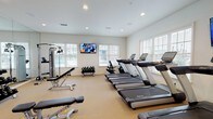 Located at 151 Du Rhu Dr in Mobile, Alabama, this well-appointed fitness center is a single-floor facility designed to support a comprehensive workout experience. The space is bright and open, with multiple large windows fitted with horizontal blinds lining the walls, allowing generous natural light to fill the room and offering views of the surrounding exterior.

The cardio section is prominently featured, housing a row of modern treadmills equipped with individual display screens, alongside a selection of elliptical cross-trainers and a stationary bike. Multiple wall-mounted flat-screen televisions are positioned throughout the room, ensuring entertainment and connectivity during workouts.

The strength training area is equally well-equipped, featuring a multi-station cable and pulley machine, a selection of selectorized weight machines, and a full rack of dumbbells ranging across various weights. Adjustable benches are available throughout the space to support both free weight and machine exercises.

A floor-to-ceiling mirror spans one wall, enhancing the sense of depth and allowing users to monitor their form during training. The flooring is a neutral carpeted surface, and recessed lighting in the white ceiling supplements the natural light. The overall ambiance is clean, functional, and inviting, making this fitness center a well-rounded amenity for residents and users of the facility.