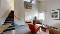 Located at 1001 Bridgeport Ave in Shelton, CT, this two-story space offers four bedrooms, two full bathrooms, and a thoughtfully designed layout spread across two floors. The main floor houses the combined kitchen and living area, and accommodates the majority of the living space, while the upper floor provides additional bedroom accommodations.

The living area immediately impresses with dramatically vaulted ceilings, a ceiling fan, and natural light streaming through multiple windows dressed in white curtains. The space flows comfortably, with neutral-toned carpeting, warm wall colors, and a staircase leading to the upper level.

The kitchen is well-appointed with white cabinetry, granite-style countertops, stainless steel appliances including a dishwasher, range, and refrigerator, as well as a pendant light fixture and a breakfast bar for casual dining.

Each of the four bedrooms features carpeted flooring, closet storage, and windows that bring in natural light. The primary bedroom is generously proportioned, offering a serene, neutral palette. Additional bedrooms include wall-mounted TVs and ample closet space.

Both bathrooms are finished with granite-top vanities, warm wood cabinetry, and tub-shower combinations. Throughout the space, vaulted and angled ceilings add architectural character, enhancing the overall sense of openness and comfort.