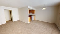 This single-floor unit is designed with a straightforward, functional layout that maximizes the use of available space. The living area is generously sized, finished with neutral-toned carpeting and a textured ceiling. A large sliding window allows natural light to fill the room, complemented by a pendant light fixture overhead. A baseboard heating system and wall-mounted air conditioning unit provide year-round climate control. The open layout connects the living space directly to the kitchen, which is configured as a galley-style room. It features warm wood cabinetry on both sides, light-toned countertops, tile flooring, and white appliances including a range, microwave, refrigerator, and dishwasher. A double-basin sink is positioned beneath upper cabinets for added convenience. The bedroom is a well-proportioned, carpeted room with a window offering outdoor views, a baseboard heater, and a flush-mount ceiling light. A second carpeted room provides additional flexible living space. The bathroom includes tile flooring, a wood-toned vanity with countertop, a full tub and shower combination, and a toilet, all presented in a clean, minimal design.