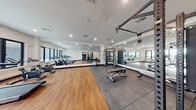 This modern fitness center offers a comprehensive workout environment designed to accommodate a wide range of exercise needs. The space is organized into distinct functional zones, each thoughtfully equipped for different training styles. The cardio area features multiple treadmills and stationary cycling bikes positioned along large, black-framed windows that flood the room with natural light and provide views of the surrounding neighborhood.

The strength training zone transitions to durable rubber flooring, housing a multi-functional cable machine, an adjustable weight bench, and a free-standing power rack stocked with a full set of dumbbells and kettlebells in graduated weights. A stability ball rounds out the functional training offerings.

Full-length mirrors line an entire wall, visually expanding the space while allowing users to monitor their form during workouts. Recessed lighting throughout the ceiling provides bright, even illumination. The main floor area features warm hardwood-style flooring, creating a clean and inviting aesthetic that contrasts with the rubberized zones. White walls and a crisp, neutral palette contribute to the bright, open ambiance, giving the fitness center a polished, contemporary feel overall.