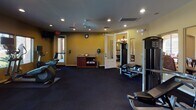 This fitness center offers a comprehensive workout environment designed to support a wide range of exercise routines. The space is laid out across a single floor and is finished with dark rubberized flooring throughout, providing a durable and functional surface suited to both cardio and strength training activities.

Along one wall, a row of treadmills and cardio machines is positioned in front of large windows fitted with horizontal blinds, allowing natural light to filter in while offering views of the surrounding outdoor greenery. Each cardio unit features an individual monitor, enhancing the workout experience.

The main floor area houses a diverse selection of strength training equipment, including a cable machine, adjustable benches, and a full rack of dumbbells ranging across multiple weights. A ceiling fan is mounted overhead, contributing to air circulation throughout the room. Recessed lighting provides consistent illumination, complemented by the natural light from the windows.

A full-length wall mirror spans a significant portion of the room, visually expanding the space and allowing users to monitor their form during workouts. A small storage console along one wall holds towels and other amenities. French doors connect the fitness area to an adjacent room, which features a chandelier, suggesting a transition to a more formal communal space nearby. The overall ambiance is practical and well-maintained, making it a fully functional facility for daily use.