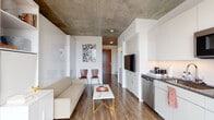Located at 1815 Division Street in Nashville's vibrant Midtown neighborhood, this single-floor studio showcases a thoughtfully designed urban living space that blends industrial and contemporary aesthetics. The most striking architectural feature is the exposed concrete ceiling, which spans the entire unit and adds an industrial character that contrasts beautifully with the clean white walls and warm wood-tone flooring throughout.

The open-concept layout maximizes functionality through smart design. A built-in Murphy bed system integrated with shelving and cabinetry serves as the room's central organizing element, allowing the living area to transition seamlessly between uses. The living space accommodates a sofa, coffee table, and additional seating, while floor-to-ceiling windows flood the space with natural light and offer sweeping views of the Nashville skyline.

The compact kitchen is efficiently outfitted with stainless steel appliances, including a refrigerator, dishwasher, and over-the-range microwave, complemented by white cabinetry, dark countertops, and a tiled backsplash. A wall-mounted flat-screen television is integrated into the cabinetry adjacent to the kitchen.

The bathroom features large-format tile flooring, a contemporary floating vanity with a wood-grain finish and open shelf, a framed mirror, and clean white walls, completing the modern aesthetic of the space.