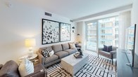 Located at 1201 Half St SE in Washington, DC's vibrant Capitol Riverfront neighborhood, this thoughtfully designed residence offers a refined modern aesthetic throughout its single-floor layout. The open-concept living area is bright and welcoming, featuring floor-to-ceiling windows that frame urban views and fill the space with natural light. A comfortable seating arrangement anchors the room, complemented by bold wall art and light hardwood flooring that flows throughout.

The kitchen is a standout feature, outfitted with sleek matte cabinetry in charcoal and soft blue tones, stainless steel appliances including a gas range and built-in oven, light quartz countertops, and a peninsula with bar seating — ideal for both everyday meals and entertaining.

The primary bedroom offers a serene retreat with a tufted upholstered headboard, dual nightstands, and warm ambient lighting. The en-suite bathroom features a double-sink vanity with a wide illuminated mirror and clean, neutral finishes. A second bedroom enjoys city views through a generous window and is styled with wall-mounted lighting and crisp, neutral tones. The second bathroom includes a soaking tub with shower, floating vanity, and backlit mirror, maintaining the home's consistent modern palette.