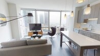 This stylish high-rise residence offers a thoughtfully designed open-concept layout that seamlessly connects the living, dining, and kitchen areas. Floor-to-ceiling windows flood the main living space with natural light and frame dramatic panoramic views of the surrounding city skyline and waterfront beyond — a defining feature of the home. The living area is anchored by dark hardwood-style flooring and accommodates both a comfortable seating arrangement and a sleek dining setup, all bathed in natural light.

The kitchen is outfitted with flat-front, wood-toned cabinetry extending to the ceiling, a white quartz countertop, a marble-style backsplash, and a full suite of stainless steel appliances including a gas range, built-in microwave, and French door refrigerator. Pendant lighting adds a warm, refined touch above the kitchen island and dining area.

The bedroom is a serene retreat, featuring large windows with city views, plush carpeting, and a dedicated walk-in closet offering generous storage. The bathroom is clean and contemporary, presenting a light wood vanity with a white quartz countertop, a frameless glass walk-in shower with large-format white tile, and dark tile flooring. Every detail reflects a cohesive modern aesthetic designed for comfortable urban living.