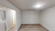 This single-floor unit offers a straightforward, well-maintained layout designed for comfortable everyday living. Consistent light wood-style laminate flooring runs throughout the space, complemented by neutral gray-beige walls and clean white baseboards, creating a cohesive and move-in-ready aesthetic.

The living area is open and adequately sized, receiving natural light through windows fitted with white horizontal blinds. A wall-mounted PTAC unit provides climate control, and a front entry door with a side window adds both accessibility and additional natural light to the main living space.

The kitchen is efficiently arranged in an L-shaped configuration, featuring white shaker-style cabinetry offering generous storage above and below the counters. A granite-look laminate countertop ties the space together, accompanied by a double stainless steel sink and a white electric coil range with an under-cabinet ventilation hood. A white refrigerator completes the appliance package.

The bedrooms are comfortably proportioned, each equipped with a flush-mount ceiling light fixture and windows that allow natural views of surrounding greenery. The bathroom includes a full tub and shower combination with a white fiberglass surround. Overall, the unit presents a clean, functional environment with consistent finishes throughout.