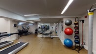 This fitness center is a thoughtfully designed, single-floor facility offering a well-rounded selection of exercise equipment to support a variety of workout routines. The space features durable rubber tile flooring throughout, providing both safety and comfort underfoot. Recessed linear LED lighting runs across the ceiling, ensuring bright, even illumination across the entire facility.

The cardio zone is anchored by multiple True-brand treadmills with integrated touchscreen displays, alongside elliptical trainers and stationary bikes — including a True upright cycle — all equipped with modern digital consoles. A dedicated strength area includes a cable resistance machine and an adjustable workout bench, catering to functional and resistance training needs.

Full-length wall mirrors are strategically positioned to visually expand the space and allow users to monitor their form during exercise. Along one wall, a vertical ball storage rack holds a variety of stability and medicine balls in multiple sizes, complemented by resistance bands — making the space well-suited for functional fitness and core training.

The clean, white-painted walls and uncluttered layout contribute to an open, focused atmosphere. A small window allows a modest amount of natural light to enter, and a sanitizing station is conveniently mounted near the entrance, reflecting a commitment to cleanliness and user well-being.