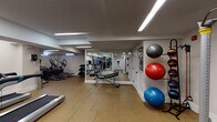 This fitness center is a thoughtfully designed, single-floor facility offering a well-rounded selection of exercise equipment to support a variety of workout routines. The space features durable rubber tile flooring throughout, providing both safety and comfort underfoot. Recessed linear LED lighting runs across the ceiling, ensuring bright, even illumination across the entire facility.

The cardio zone is anchored by multiple True-brand treadmills with integrated touchscreen displays, alongside elliptical trainers and stationary bikes — including a True upright cycle — all equipped with modern digital consoles. A dedicated strength area includes a cable resistance machine and an adjustable workout bench, catering to functional and resistance training needs.

Full-length wall mirrors are strategically positioned to visually expand the space and allow users to monitor their form during exercise. Along one wall, a vertical ball storage rack holds a variety of stability and medicine balls in multiple sizes, complemented by resistance bands — making the space well-suited for functional fitness and core training.

The clean, white-painted walls and uncluttered layout contribute to an open, focused atmosphere. A small window allows a modest amount of natural light to enter, and a sanitizing station is conveniently mounted near the entrance, reflecting a commitment to cleanliness and user well-being.