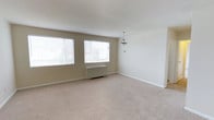 This single-floor residence offers a clean, functional layout designed for comfortable everyday living. The main living area immediately draws attention with its generous proportions, soft wall-to-wall carpeting in a neutral beige tone, and crisp white ceilings that contribute to an open, airy feel. Two large windows fitted with horizontal blinds flood the room with abundant natural light, creating a bright and welcoming atmosphere throughout the day. The walls are finished in a warm, understated beige, providing a versatile backdrop for a variety of interior styles. A classic pendant light fixture is centrally positioned, adding a simple yet elegant focal point to the space. A baseboard heating unit beneath the windows ensures year-round comfort. White baseboards throughout offer clean, defined transitions between the walls and flooring, lending the space a tidy, well-maintained appearance. The overall ambiance is calm and neutral, making it an ideal canvas for personalization. The single-floor layout promotes ease of movement and accessibility, with a straightforward, uncluttered design that prioritizes practicality and livability.