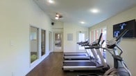 This fitness center is designed to provide a functional and comfortable workout environment within a single-floor layout. The space features a row of treadmills and cardio machines positioned along the windowed wall, allowing exercisers to enjoy natural light and outdoor views during their workouts. Large windows line the exterior wall, flooding the room with daylight and creating an open, airy atmosphere. The floors are finished in rich, dark hardwood, offering both durability and a clean aesthetic. A ceiling fan is mounted overhead, complementing the recessed lighting to ensure consistent airflow and visibility throughout the space. The walls are painted in a neutral, light tone that enhances the sense of openness. Interior glass partitions and doorways provide views into adjacent areas, suggesting additional amenity spaces nearby, including what appears to be a yoga or stretching room with free weights. An informational board is mounted on the wall, offering guidance for facility users. Overall, the fitness center presents a well-organized, inviting space suited for daily cardiovascular and general exercise routines.