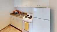 This junior one-bedroom, one-bathroom unit is laid out efficiently across a single floor, offering a practical and straightforward living space. The overall design is clean and neutral, with white walls throughout that create a bright, open feel.

The main living area is carpeted in a light beige tone and receives natural light through a window fitted with horizontal blinds, lending the space a welcoming atmosphere. The area is open enough to accommodate standard living room furnishings comfortably.

The kitchen is compact and well-appointed, featuring white cabinetry with rounded hardware above and below the counter, granite-style laminate countertops, a stainless steel sink, and white appliances including a coil-burner electric range and a full-sized refrigerator. The flooring transitions to tile in this area, providing a practical and easy-to-clean surface.

The bedroom is carpeted and finished with neutral white walls. A mirrored sliding-door closet provides storage and visually expands the room, with interior shelving and a hanging rod visible behind the panels.

The bathroom features a full-sized soaking tub with a handheld showerhead, chrome fixtures, and white square tile surround, maintaining a clean and functional aesthetic consistent with the rest of the unit.