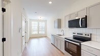 Located at 700 E. Long St. in Columbus, Ohio, this well-designed one-bedroom, two-bathroom unit offers a thoughtful layout that maximizes every square foot of its compact footprint. The kitchen is a standout feature, appointed with gray shaker-style cabinetry, white subway tile backsplash, light quartz countertops, and a full suite of stainless steel appliances including a range, microwave, dishwasher, and refrigerator. Light hardwood flooring runs through the main living and kitchen areas, lending a warm, contemporary feel. The open sightline from the kitchen toward the living area creates an airy sense of flow, with natural light streaming in through large windows fitted with horizontal blinds. The bedroom is carpeted for comfort and features multiple windows with urban views, filling the room with generous natural light. A dedicated laundry area includes a stacked washer and dryer unit alongside a built-in closet with hanging rod — a practical and space-efficient amenity. The bathrooms are clean and modern, with one featuring a teal vanity with matte black hardware accents and hardwood-style flooring. The overall aesthetic is bright and neutral, with white walls and crisp finishes throughout.
