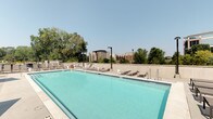 This outdoor amenity space centers around a generously sized lap pool with clear, turquoise water set against light-toned pool decking. The pool is well-defined with marked depth indicators and equipped with multiple entry points via pool ladders. Surrounding the pool on multiple sides is a broad sundeck furnished with rows of cushioned lounge chairs, providing ample space for relaxation and sunbathing. Tall pole-mounted light fixtures are positioned throughout the deck, ensuring the area remains well-lit during evening hours. A low perimeter wall encloses the space, offering both safety and a clean, modern aesthetic while maintaining open sightlines. Beyond the wall, mature deciduous trees create a natural green backdrop, softening the urban environment. Views of nearby mid-rise and multi-story residential buildings are visible in the background beneath a clear, open sky, giving the space a connected yet private feel. The overall design emphasizes functionality and comfort, making it well-suited for both active recreation and leisure. The combination of ample seating, a full-length pool, and open-air surroundings creates an inviting and well-appointed outdoor retreat.