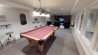 Located at 4215 S 30th St in Tacoma, Washington, this single-floor recreation space is designed with entertainment and leisure in mind. The room is anchored by a full-size pool table with a rich red felt surface and warm wood frame, illuminated by an elegant wrought-iron pendant light fixture with frosted glass shades that adds a classic touch to the space. The carpeted flooring and neutral wall tones create a comfortable, inviting atmosphere throughout. Toward the back of the room, a dedicated home theater area features multiple reclining leather chairs and a large flat-screen television, making it ideal for movie nights or gaming. Built-in wooden shelving units flank the media area, providing ample storage and display space for media, books, and accessories. Recessed ceiling lighting supplements the pendant fixture, ensuring the entire room is well-lit. Decorative wall-mounted game boards and shelving accents add personality and a playful character to the space. The generous open layout allows for easy movement between the billiards and lounge areas, making this a versatile and well-appointed recreation room suited for hosting and relaxation alike.