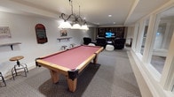 Located at 4215 S 30th St in Tacoma, Washington, this single-floor recreation space is designed with entertainment and leisure in mind. The room is anchored by a full-size pool table with a rich red felt surface and warm wood frame, illuminated by an elegant wrought-iron pendant light fixture with frosted glass shades that adds a classic touch to the space. The carpeted flooring and neutral wall tones create a comfortable, inviting atmosphere throughout. Toward the back of the room, a dedicated home theater area features multiple reclining leather chairs and a large flat-screen television, making it ideal for movie nights or gaming. Built-in wooden shelving units flank the media area, providing ample storage and display space for media, books, and accessories. Recessed ceiling lighting supplements the pendant fixture, ensuring the entire room is well-lit. Decorative wall-mounted game boards and shelving accents add personality and a playful character to the space. The generous open layout allows for easy movement between the billiards and lounge areas, making this a versatile and well-appointed recreation room suited for hosting and relaxation alike.
