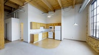 This 2-bedroom, 2-bathroom unit offers a distinctive industrial-loft aesthetic on a single level. The most striking architectural feature is the dramatic vaulted ceiling adorned with exposed wooden beams and natural wood paneling, which lends the space an open, airy feel despite its compact footprint. A ceiling fan is centrally positioned to complement the generous ceiling height, while pendant lighting adds a modern touch to the living area.

The kitchen is thoughtfully designed in an L-shaped configuration, featuring warm honey-toned wood cabinetry, a white appliance suite including a refrigerator, electric range, and built-in microwave, as well as a double basin sink. The kitchen transitions seamlessly into the living area via a peninsula that provides additional counter space and casual seating potential. Hardwood-style flooring defines the kitchen zone, while plush carpeting covers the main living area, creating a clear and comfortable delineation between the two spaces.

The open-plan layout maximizes the usable square footage, allowing natural sightlines across the full width of the unit. The white walls provide a clean, neutral backdrop that complements the warm tones of the wood elements throughout, making the space feel bright and welcoming.