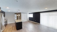 This single-floor space showcases an open-concept layout that seamlessly connects the kitchen and living areas, creating a cohesive and functional environment. The kitchen features a dark-cabineted island topped with granite countertops, illuminated by stylish pendant lights that add a modern touch. The living area is defined by a striking navy blue accent wall, which provides a bold contrast against the neutral grey tones found throughout the rest of the space.

Durable luxury vinyl plank flooring extends across the entire main area, offering a clean and contemporary aesthetic that is both practical and visually appealing. The textured ceiling adds a traditional finish overhead. Natural light enters through a window on the accent wall side, complemented by vertical blinds on a larger sliding door or window, offering privacy and light control.

A hallway visible in the background leads to additional areas of the unit, while a set of white double doors provides access to a closet, adding ample storage. The overall ambiance is modern and well-appointed, with a thoughtful contrast between dark and light design elements throughout.