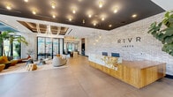 Located at 307 SW 5th St in Fort Lauderdale, FL, the RIVR Lofts lobby is a generously sized, single-floor communal space that makes a bold first impression through its contemporary design and thoughtful detailing. The entrance area is anchored by a dramatic reception desk finished in warm wood and veined marble, set against a full-length whitewashed brick accent wall bearing the RIVR Lofts logo — a refined yet industrial aesthetic that sets the tone for the entire space.

The main lobby opens into a bright, airy lounge bathed in natural light from expansive floor-to-ceiling windows that line the exterior walls, offering views of lush outdoor greenery. A curated mix of sculptural seating in warm mustard and teal tones is arranged across patterned tile and area rug zones, creating distinct yet connected social areas. A sculptural pendant chandelier adds a dramatic focal point overhead.

Adjacent to the lounge, a sleek bar counter with dark stone surfaces and bar-height seating is framed by a striking wall installation of suspended globe pendants and lush moss spheres set into warm wood shelving — a standout design feature. The dark navy ceiling throughout contrasts beautifully with the light polished concrete floors, while abundant greenery and potted plants reinforce a vibrant, nature-connected atmosphere throughout this expansive communal space.