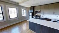 This single-floor space presents a seamlessly integrated open-concept layout that combines the kitchen and living areas into one cohesive, inviting environment. The kitchen is a standout feature, showcasing dark, contemporary cabinetry contrasted by a crisp white quartz peninsula countertop that doubles as a casual dining or workspace surface. Stainless steel appliances — including a gas range, built-in microwave, refrigerator, and dishwasher — lend a sleek, professional quality to the space. A classic white subway tile backsplash adds a timeless yet modern touch behind the range and sink. The open layout flows naturally into the living area, where warm-toned hardwood flooring runs throughout, enhancing the sense of continuity and space. Large, double-hung windows along the exterior wall flood the interior with abundant natural light, creating a bright and airy atmosphere. Neutral wall tones complement the dark cabinetry and light flooring, striking a balanced, modern aesthetic. Structural beam details at the ceiling add subtle architectural character. Overall, the space combines functional design with modern finishes, offering a clean, well-appointed interior that is both practical and visually refined.