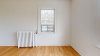 Located at 5 Donlands Ave in Toronto's East End, this one-bedroom, one-bathroom unit offers a functional and efficiently laid-out single-floor residence. The interior is finished with warm hardwood flooring throughout the main living areas and bedroom, lending a clean and inviting ambiance to the space.

The living room is bright and open, benefiting from natural light that enhances the neutral white walls and light wood floors. The bedroom is a comfortable size, featuring a classic cast-iron radiator for heating and a window that looks out to the surrounding neighbourhood, allowing for ample natural light. The hardwood flooring continues here, adding warmth and continuity.

The kitchen is finished with light ceramic tile flooring and includes a window with tree views, providing a pleasant atmosphere for cooking. Cabinetry and a full-size refrigerator complete the practical setup.

The bathroom features full white ceramic tile walls surrounding a soaking tub and shower combination, with matching tile flooring, giving it a clean and classic appearance. A hallway connects all rooms in a straightforward layout, optimizing the use of the available space.

Overall, this unit presents a tidy, no-frills design with consistent finishes, classic architectural details, and a practical configuration suited for comfortable urban living in Toronto.