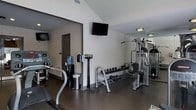 This single-floor fitness center offers a comprehensive workout environment designed to accommodate a range of exercise needs. The space is outfitted with multiple cardio machines, including treadmills and additional aerobic equipment, providing ample options for cardiovascular training. A full dumbbell rack lines one wall, offering a complete selection of free weights for strength training exercises of varying intensity. A multi-station cable and weight machine anchors one side of the room, supporting a wide array of resistance-based workouts. Wall-mounted flat-screen televisions are positioned for easy viewing from multiple points in the room, enhancing the workout experience. A water cooler is conveniently located within the space, ensuring hydration is readily accessible. Large mirrors along one wall help maximize the sense of space while allowing users to monitor their form during exercise. The flooring is finished with durable, dark rubber matting suited for high-traffic fitness use. Neutral-toned walls and recessed lighting contribute to a clean, functional ambiance throughout the facility.